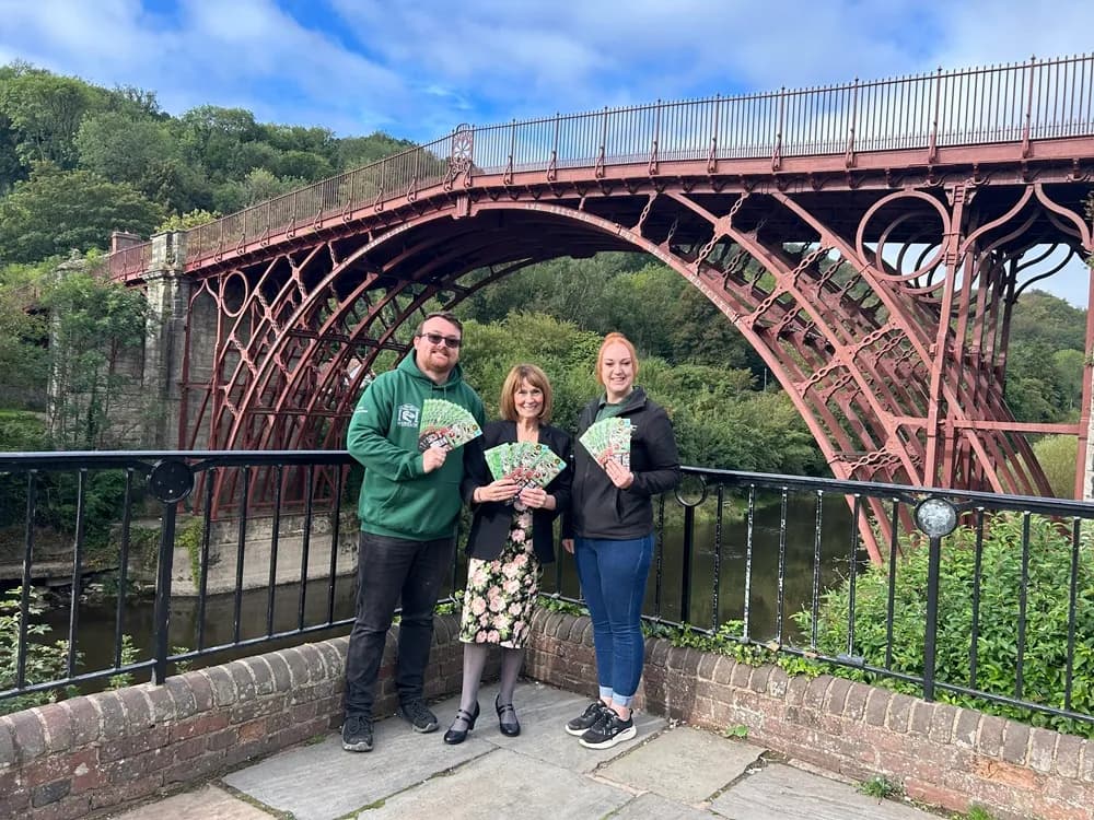 Three people stand on a paved area with a brick wall and black railing, holding green leaflets in front of the Iron Bridge over a river, surrounded by green trees under a cloudy sky.