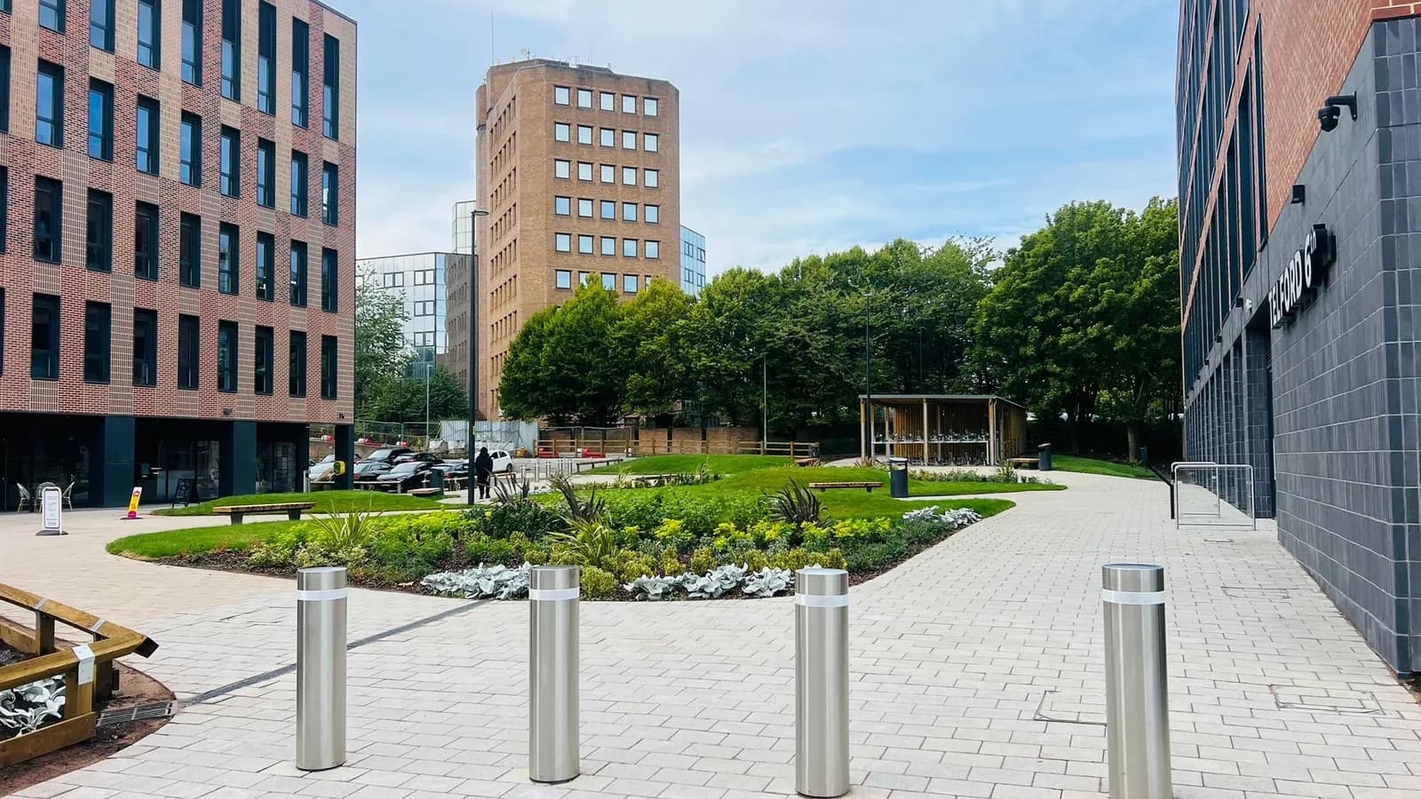 A modern paved courtyard with bollards and planters in the foreground, surrounded by multi-story buildings and trees under a partly cloudy sky.